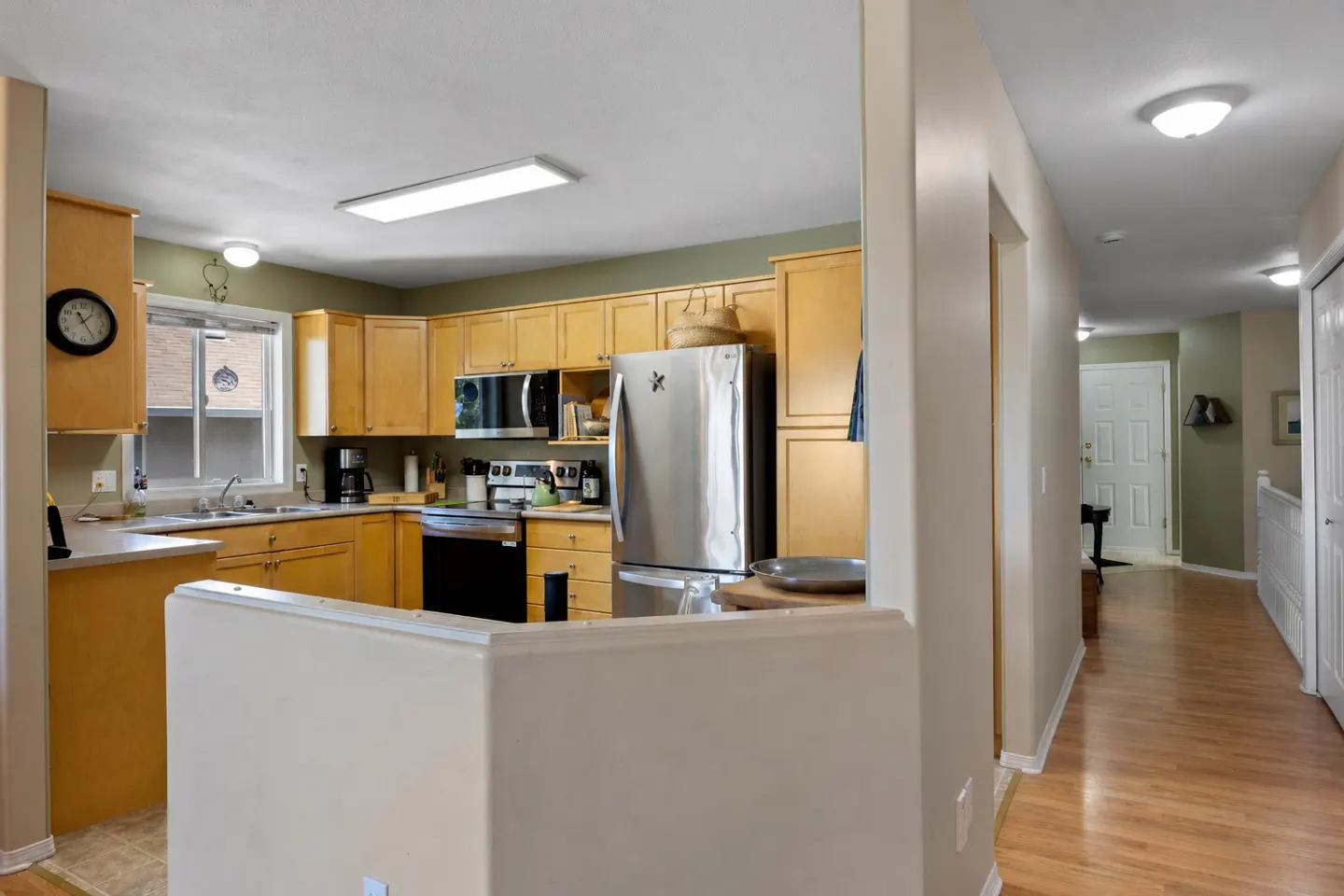 A kitchen with light wood cabinets, stainless steel appliances, and a black oven. A hallway with wood floors leads to a white door.