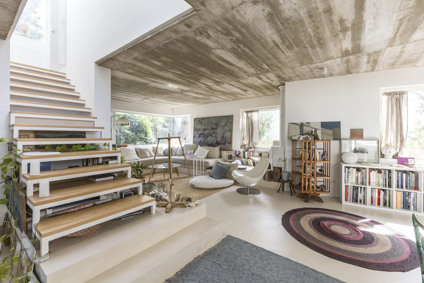 Modern living room with a floating wooden staircase, concrete ceiling, white walls, and a cozy seating area with rugs.