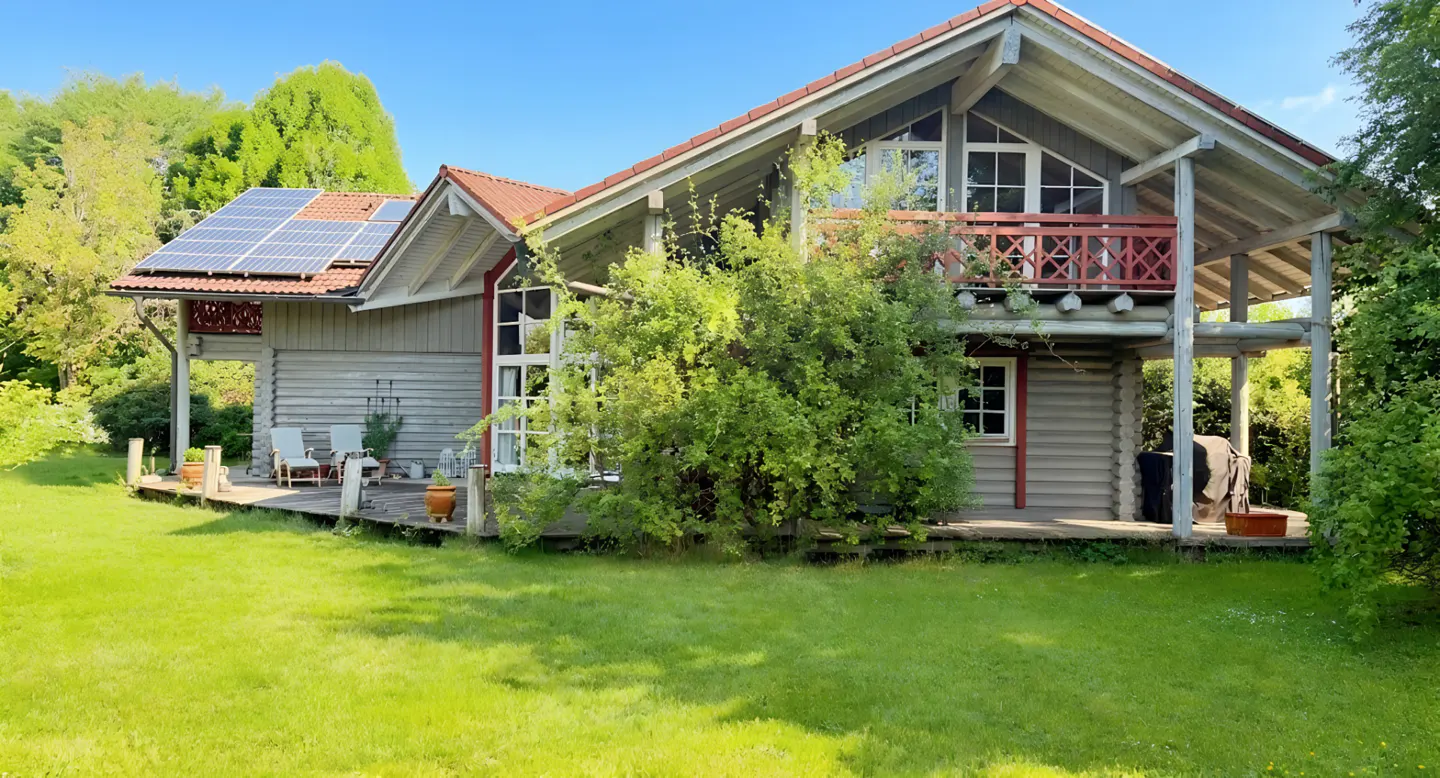 A two-story grey log cabin with a red roof, solar panels, a wooden deck, and a lush green lawn on a sunny day.