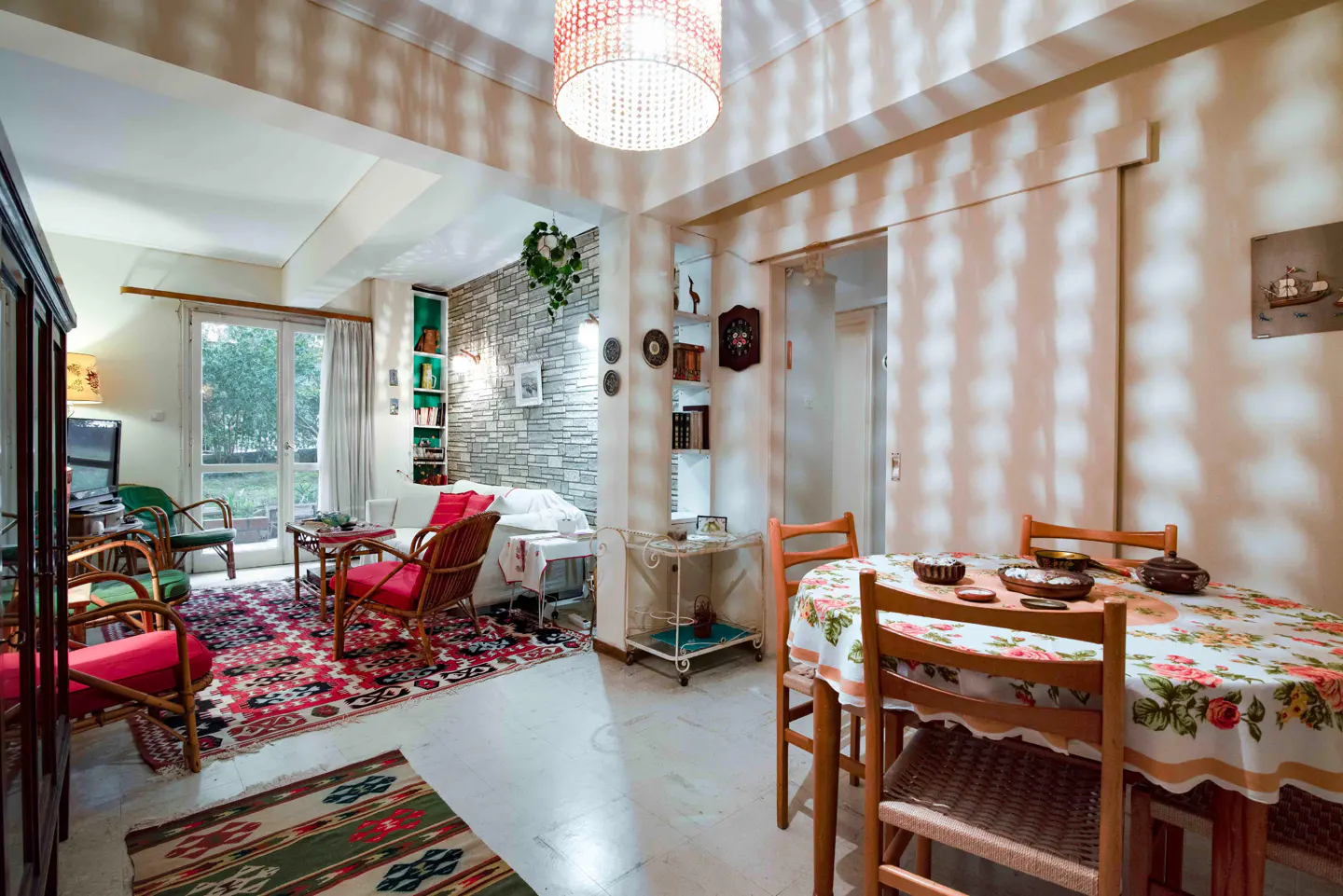 A bright living and dining area with a floral tablecloth, red wicker chairs, and a stone accent wall.