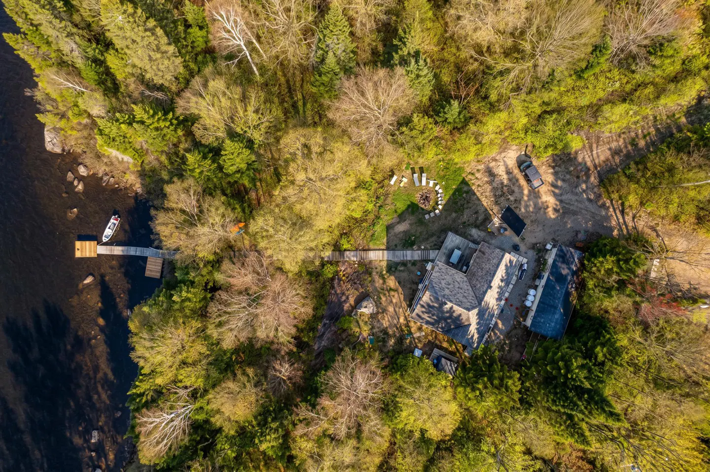Aerial view of a lake house surrounded by green trees, with a dock, boat, and fire pit.