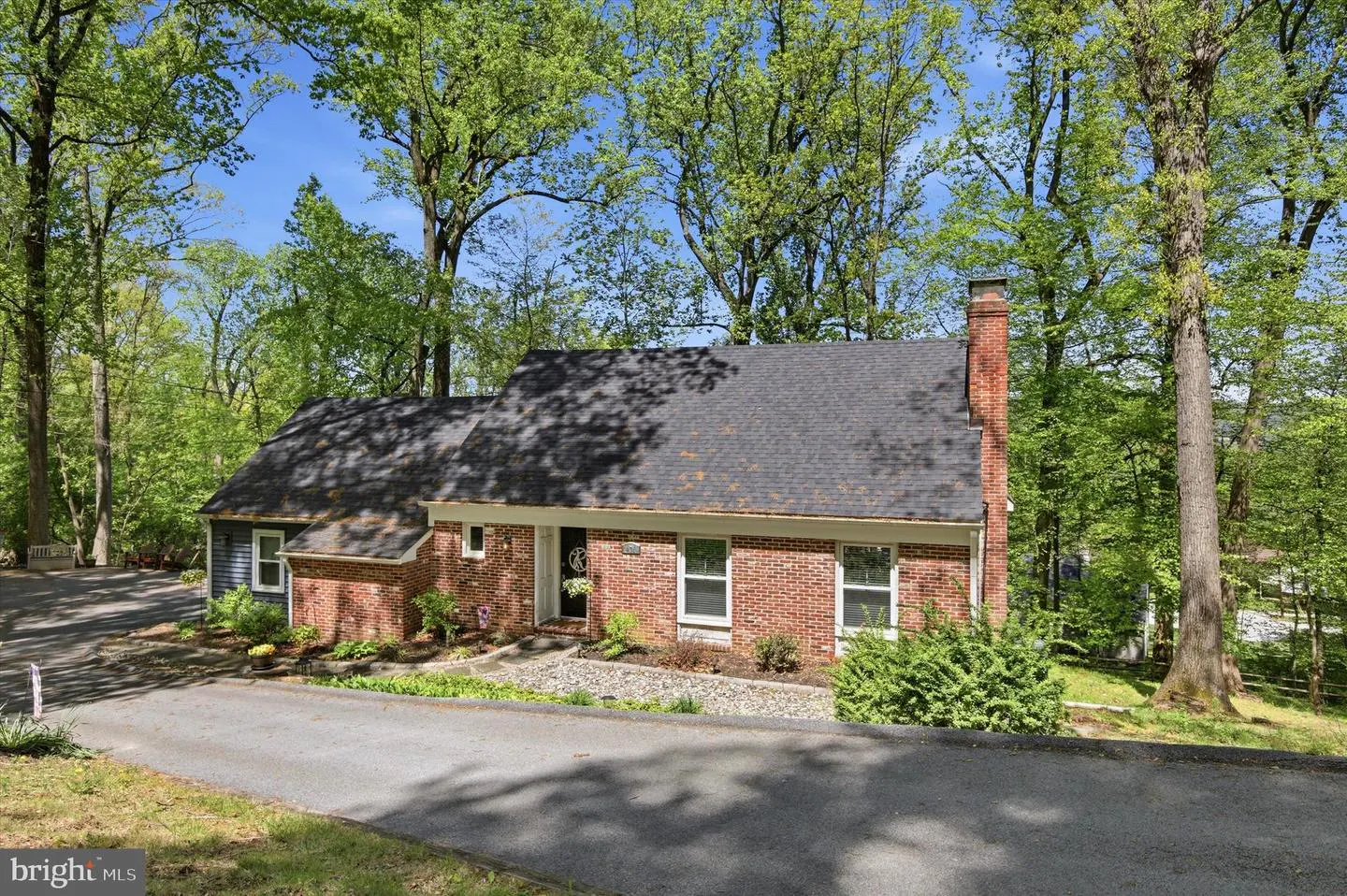 A brick house with a dark roof sits among tall green trees under a blue sky. A driveway leads up to the front of the house.