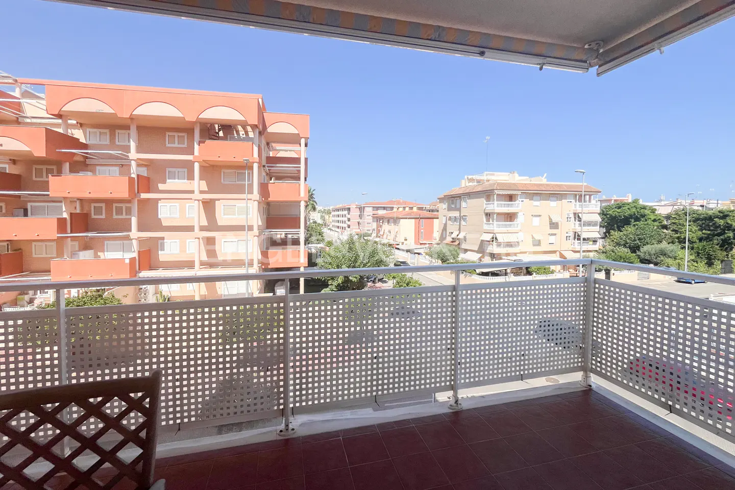 Balcony view with white lattice railing, terracotta tile floor, and a partial view of a wooden chair. Buildings and blue sky in the background.