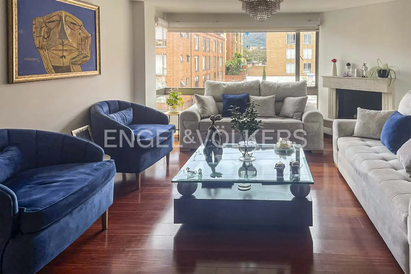 Living room with blue chairs, gray sofas, and a glass coffee table on a wood floor. A large window shows a city view.