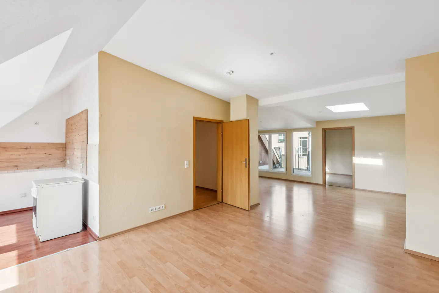 Bright, empty apartment with wood floors, beige walls, and white ceiling. A small kitchen area is visible with a white stove.
