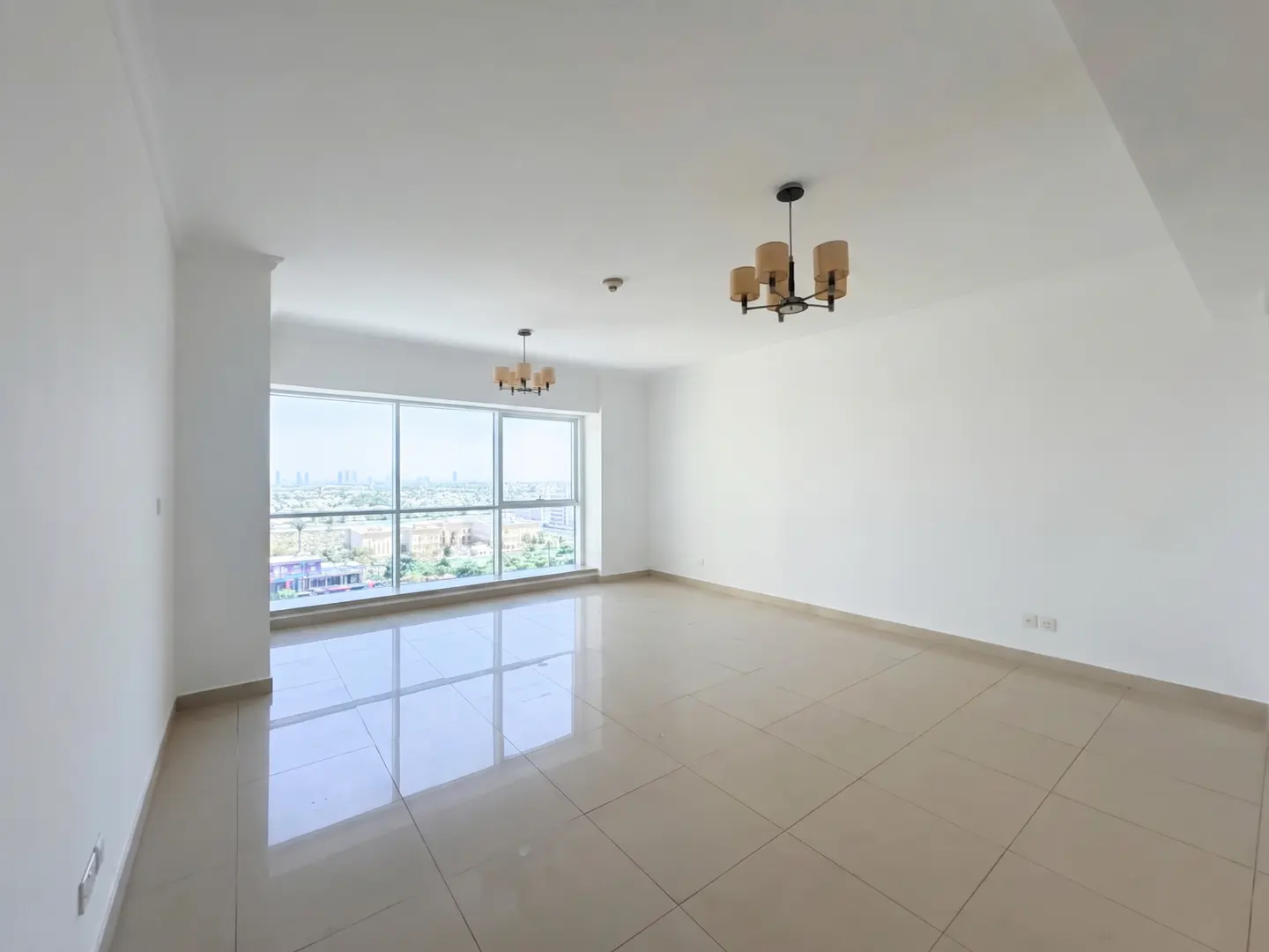 Bright, empty room with beige tile floors, white walls, and large windows showing a city view. Two chandeliers hang from the ceiling.
