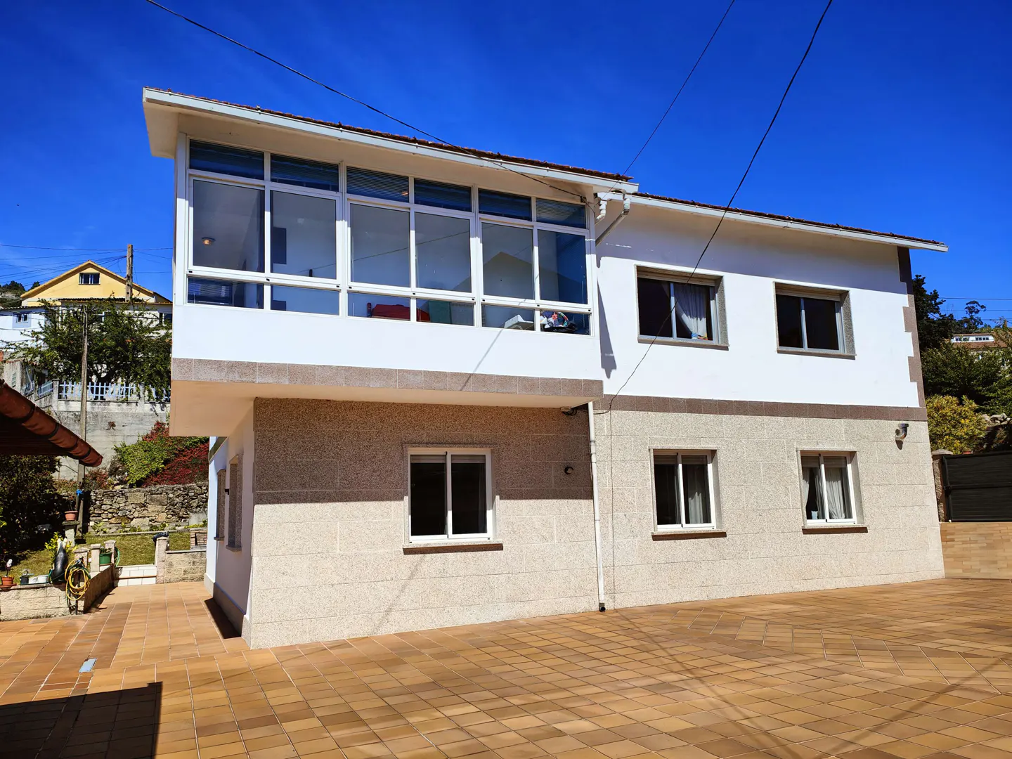 Two-story house with a stone-textured ground floor, white upper level, enclosed glass balcony, and tiled patio.