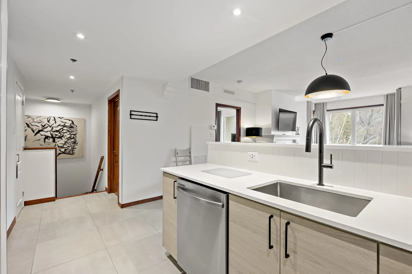 A modern kitchen with light wood cabinets, a stainless steel sink, and a black faucet. A hallway with artwork is visible in the background.