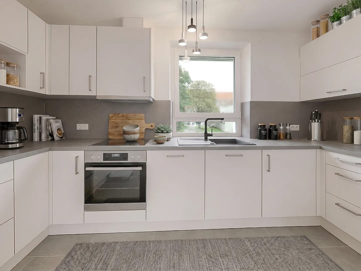 Bright kitchen with white cabinets, gray countertops, and stainless steel appliances. A window overlooks a green outdoor space.