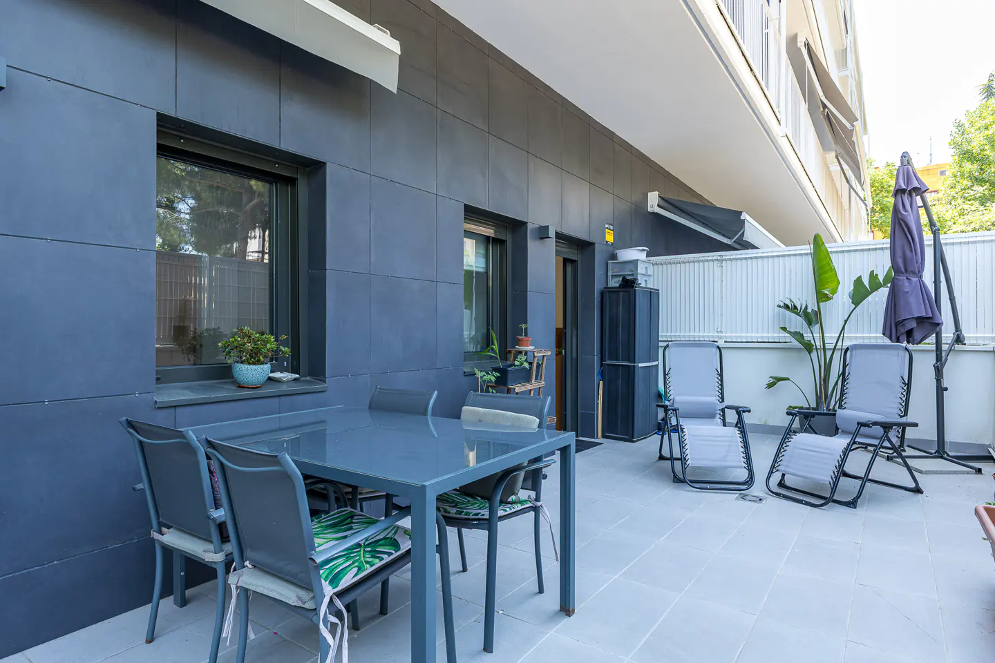 Outdoor patio with gray table, chairs, and lounge chairs. A closed umbrella and potted plants add to the ambiance.