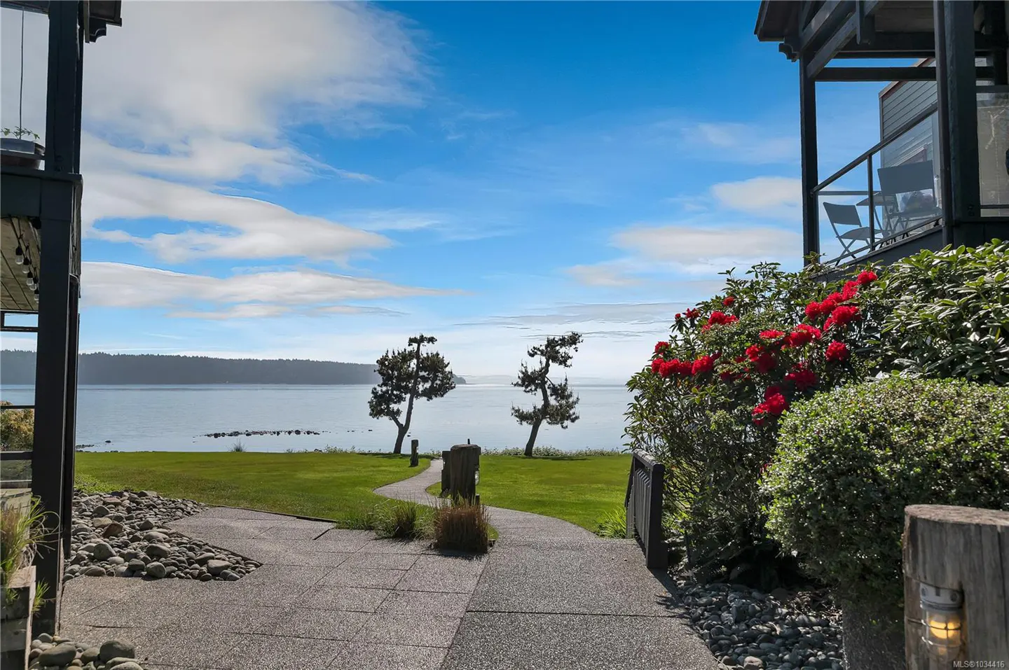 A scenic view of a waterfront property with a stone path leading to the ocean. Red flowers and green bushes frame the path. Blue sky with clouds above.