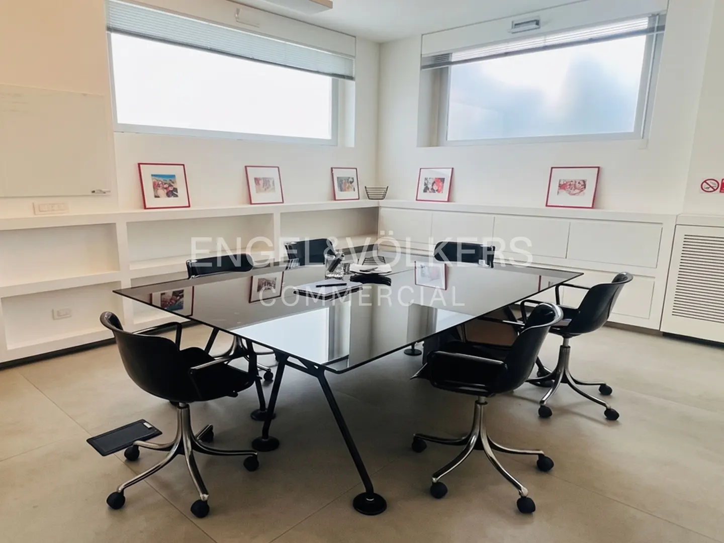 Bright office with a black conference table, black chairs, and white built-in shelves with framed pictures. Large windows provide natural light.