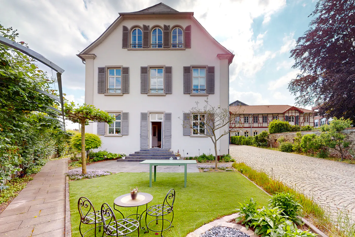 Exterior view of a three-story white house with gray shutters and a green lawn with outdoor furniture.