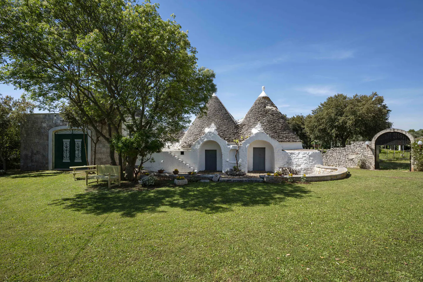 Exterior of a white Trullo house with gray conical roofs, green lawn, and a large tree.