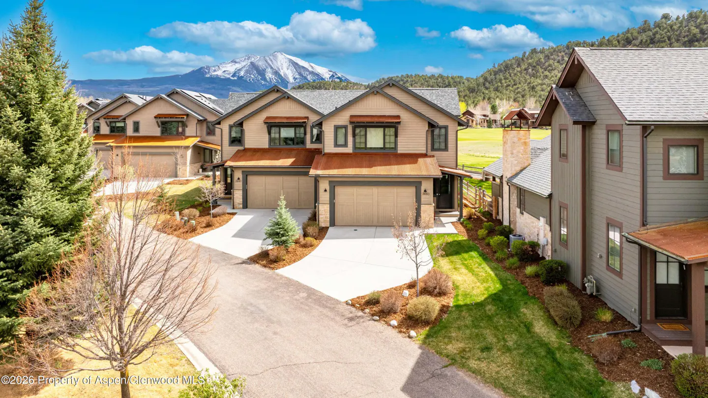 Exterior shot of tan townhouses with brown trim and garages, green grass, and a snow-capped mountain in the background.
