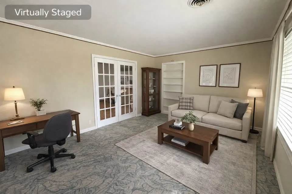 A staged living room with a desk, sofa, coffee table, and white french doors. The walls are beige, and the carpet is grey with a leaf pattern.