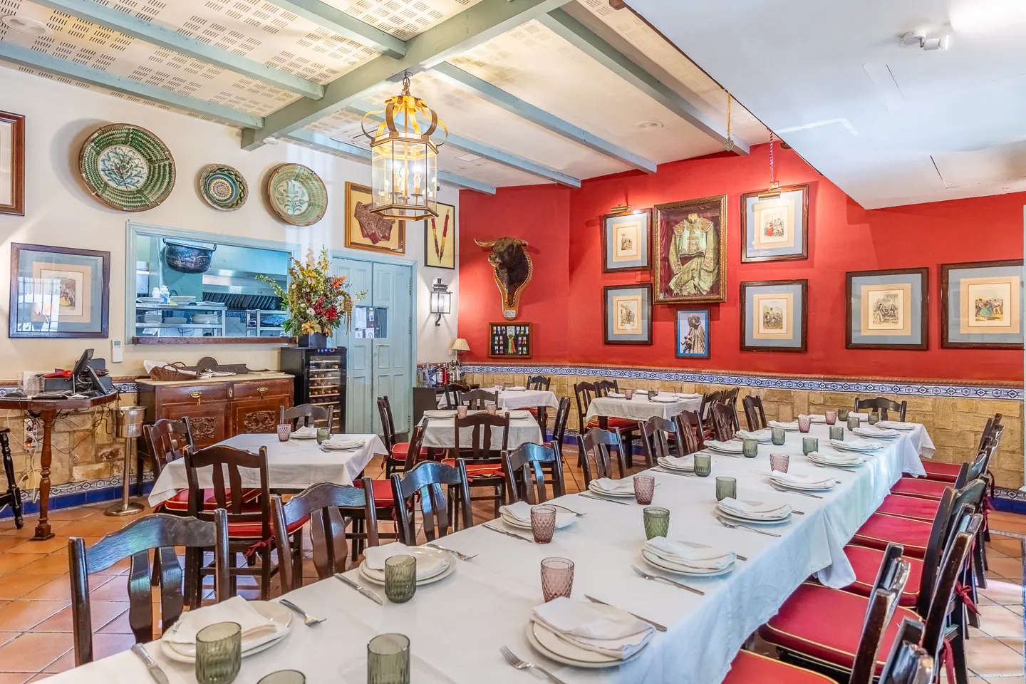 Dining room with long tables, white tablecloths, red walls, framed art, a bull head mount, and rustic wooden ceiling beams.