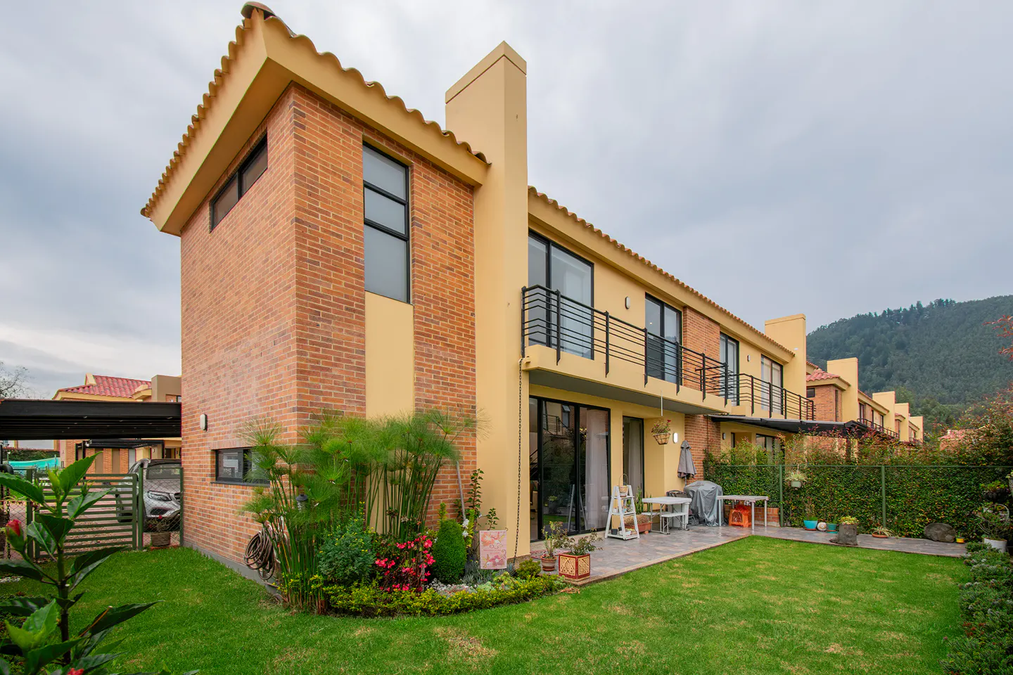Exterior view of a two-story townhouse with a brick and yellow facade, a green lawn, and a patio with outdoor furniture.