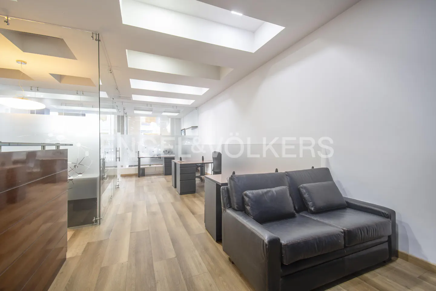 Office interior with a black leather couch, desks, and a glass-walled reception area under a skylight.