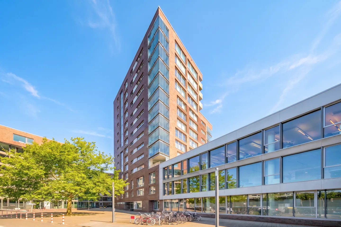 A tall, modern brown brick apartment tower stands next to a low-rise glass office building under a clear blue sky.