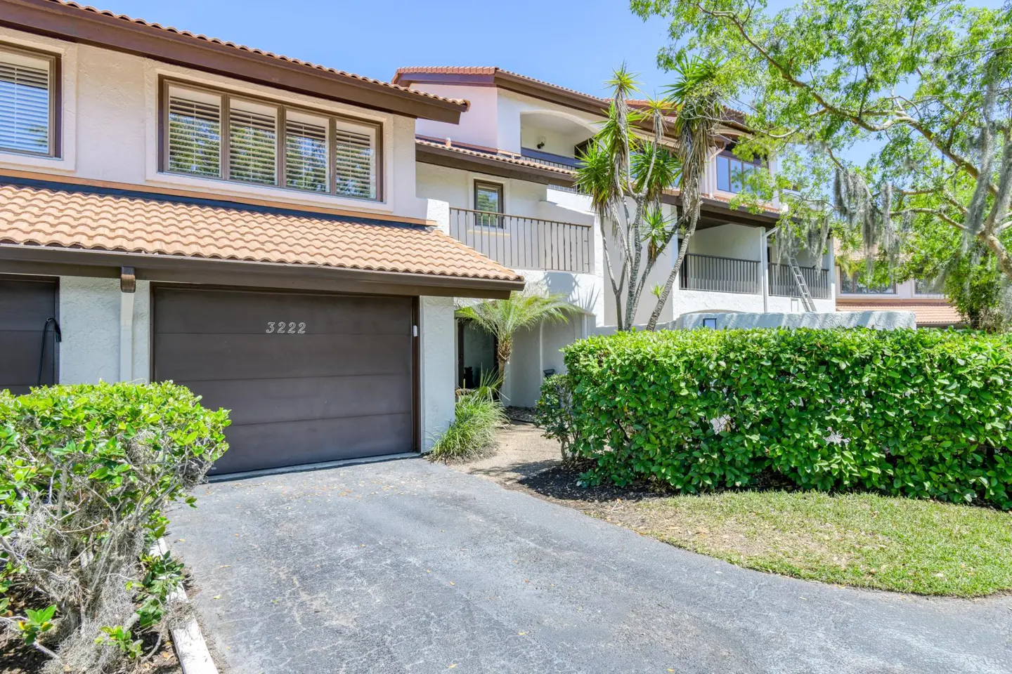 Exterior of a two-story condo with a brown garage door and tile roof. Green bushes line the driveway.