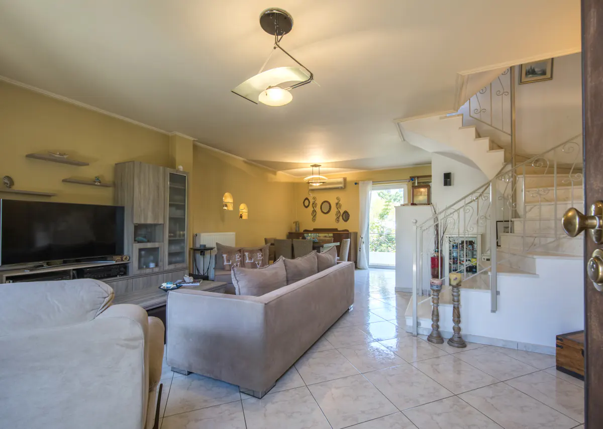 Open-plan living room with beige sofa, TV unit, tiled floors, and a staircase with white railings leading to upper level.