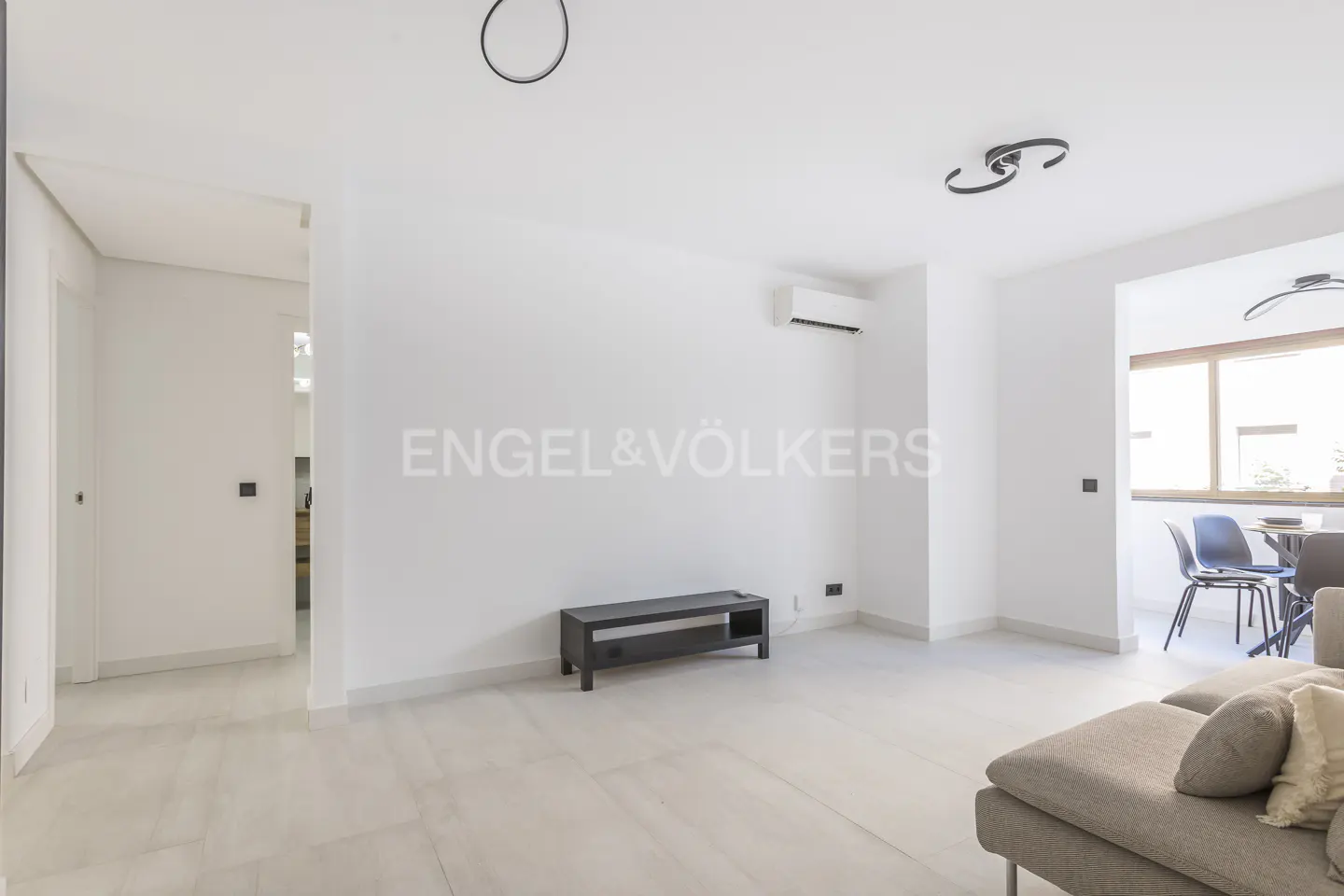 Bright, modern living room with white walls, light tile floors, and minimal decor. A gray sofa and black TV stand furnish the space. Engel & Völkers logo on the wall.