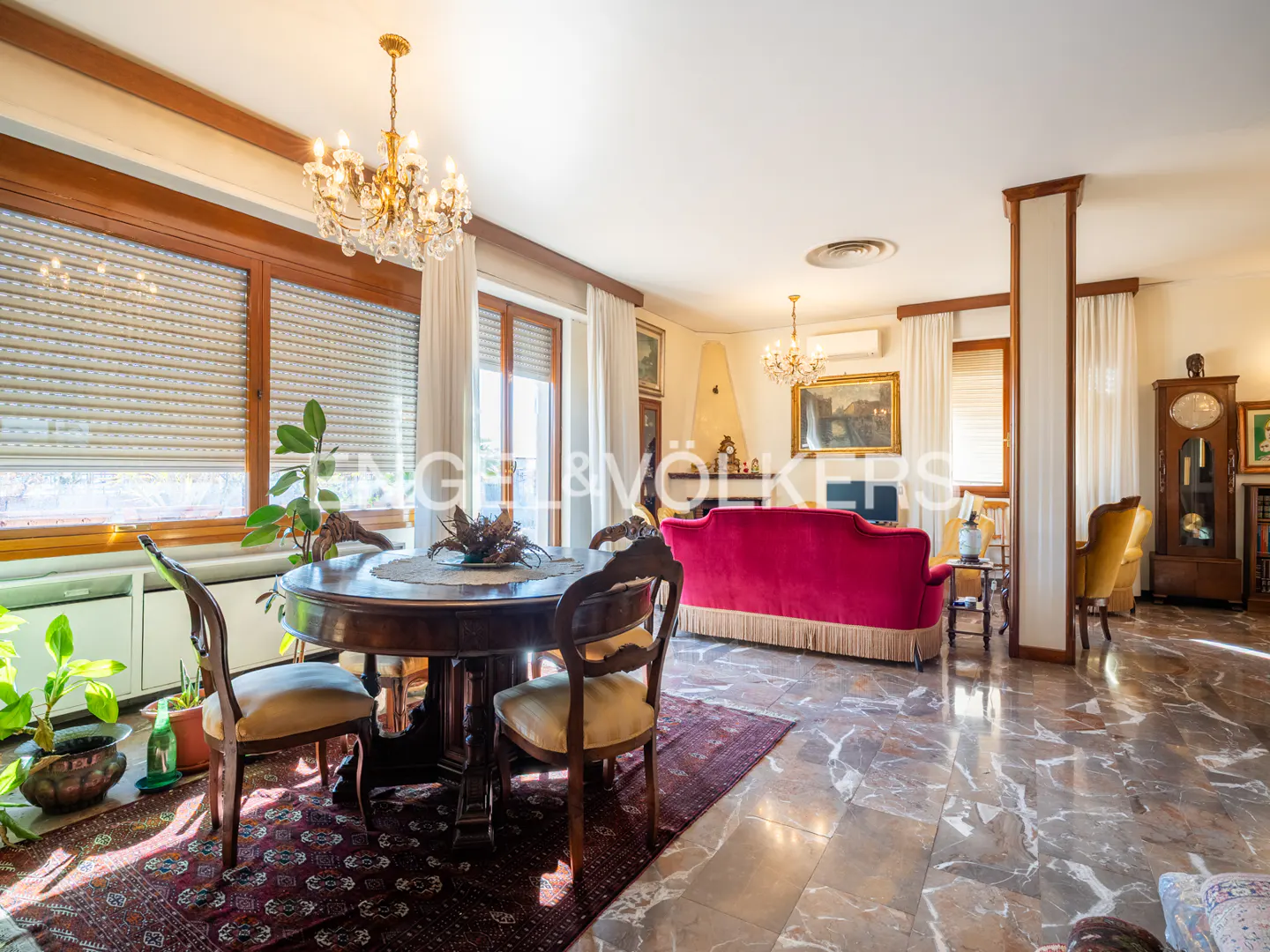 A bright living room with a round wooden table, chairs, and a red sofa. Marble floors and a chandelier add elegance.