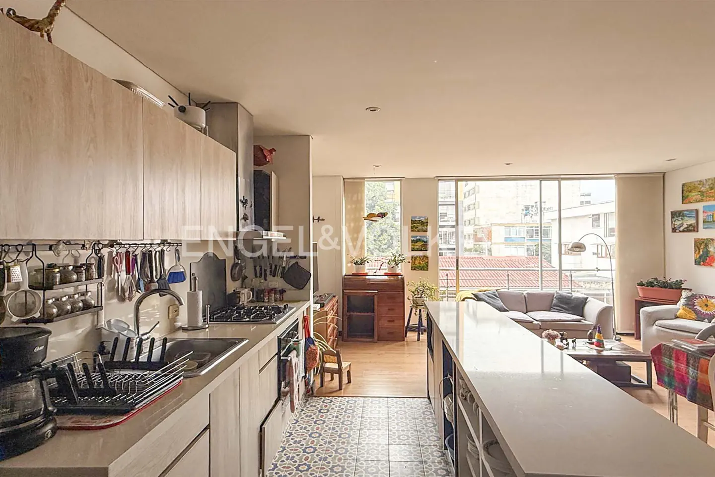 Open-concept apartment featuring a kitchen with light wood cabinets and a long island, flowing into a living room with large windows.