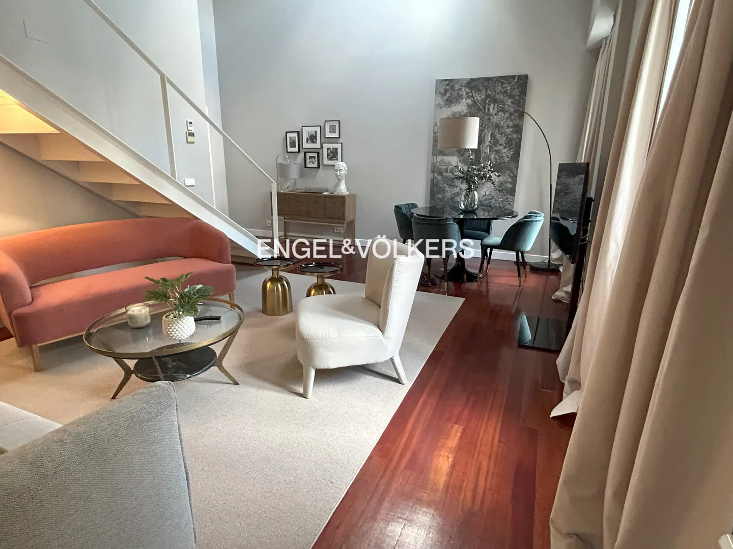 Living room with a pink sofa, white chair, round table, and dining area with blue chairs. Hardwood floors and neutral walls.