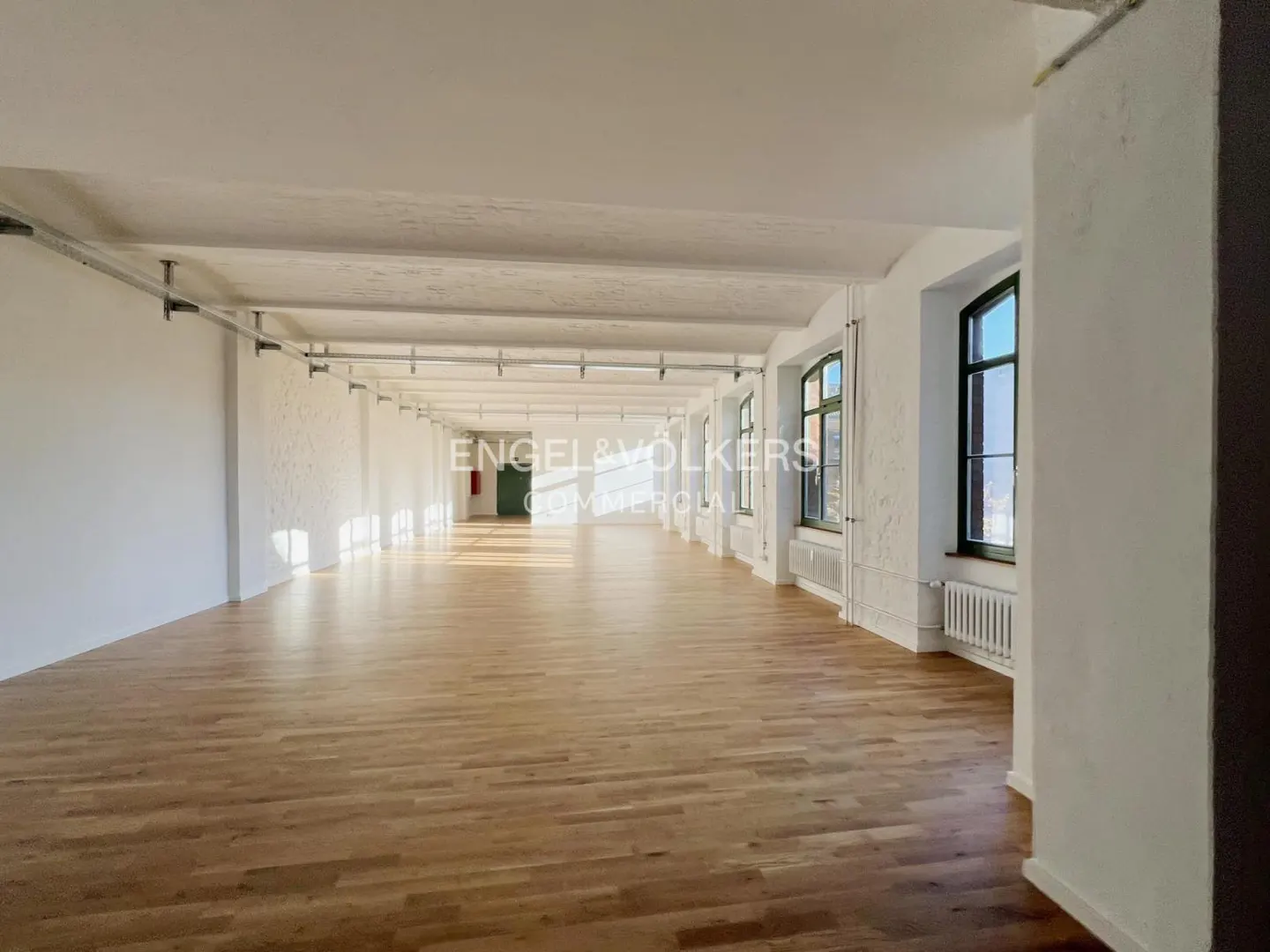 Bright, empty commercial space with hardwood floors, white walls, and large windows. Engel & Völkers logo visible in the background.