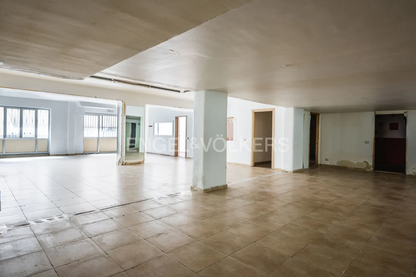 A large, empty room with beige tile floors, white walls, and multiple doorways. Natural light enters from windows on the left.