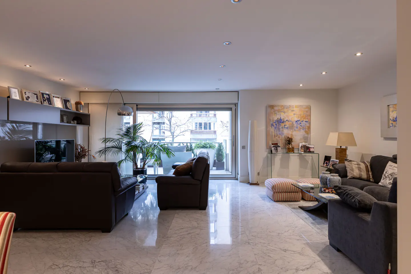 Spacious living room with marble floors, dark sofas, a large glass balcony door, and abstract wall art.