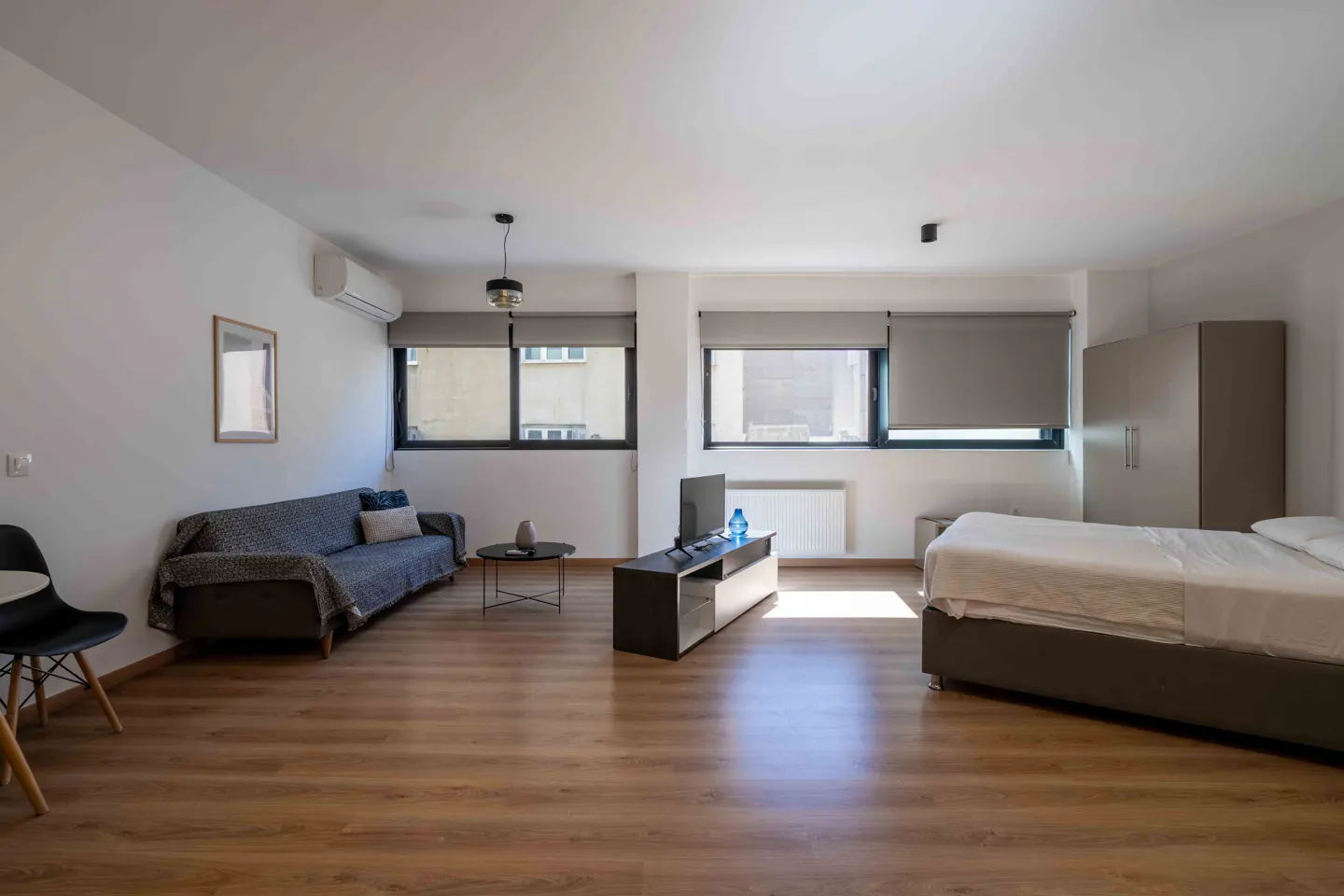 Bright studio apartment with wood floors, white walls, and large windows. A sofa, TV, bed, and wardrobe furnish the space.
