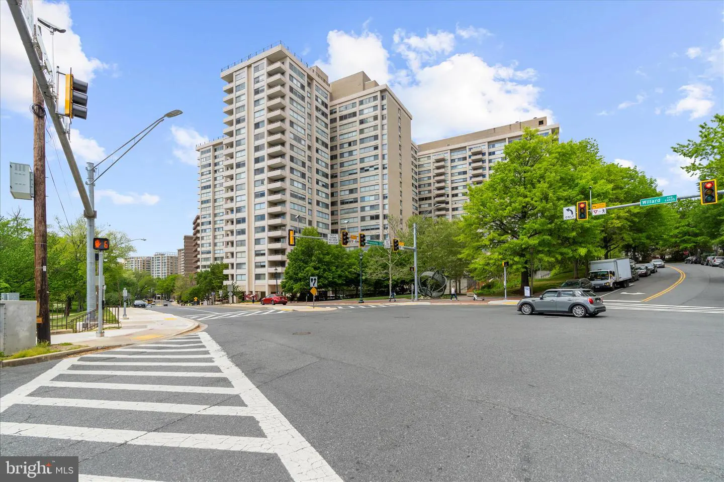 A gray car turns at a sunny intersection in front of a large, multi-story beige apartment building surrounded by trees.
