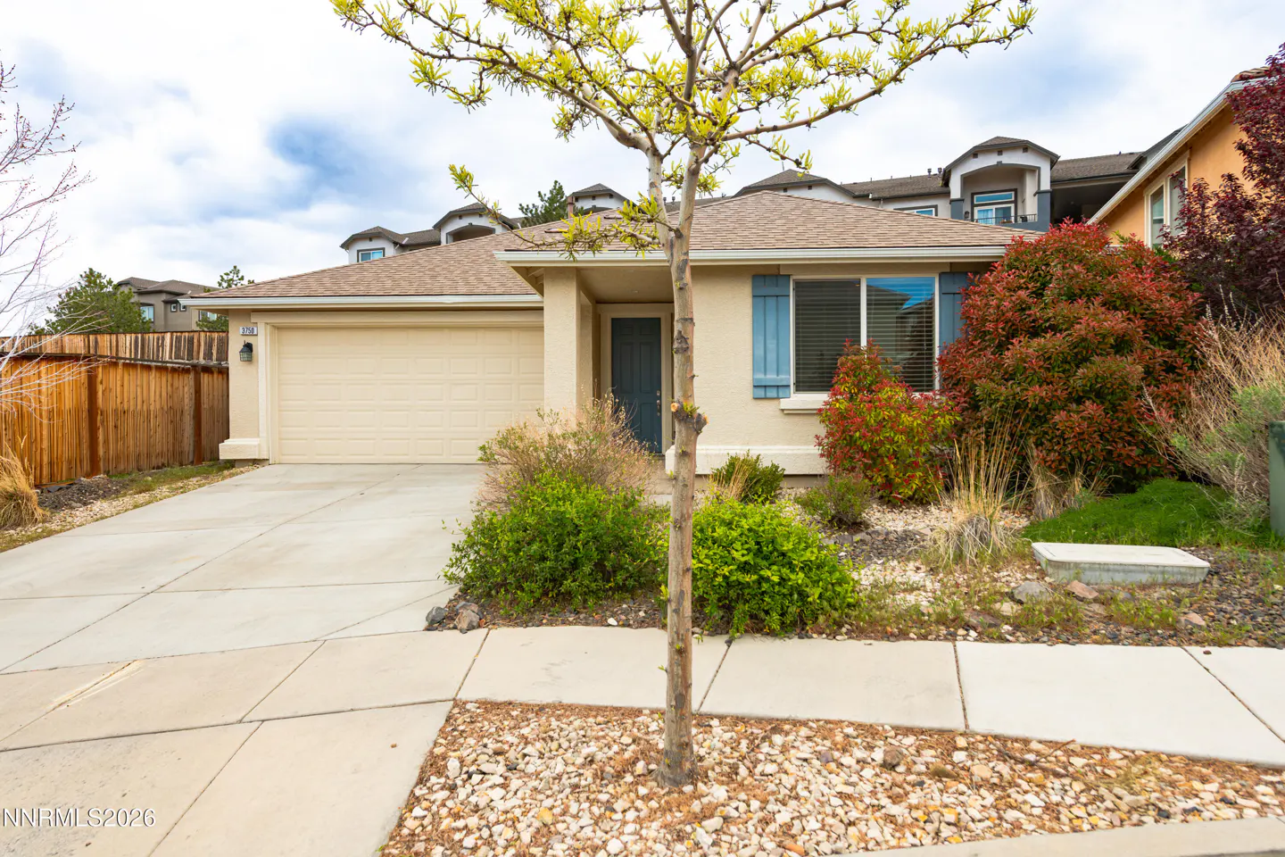Beige single-story house with a two-car garage, blue shutters, and a small tree in the front yard.