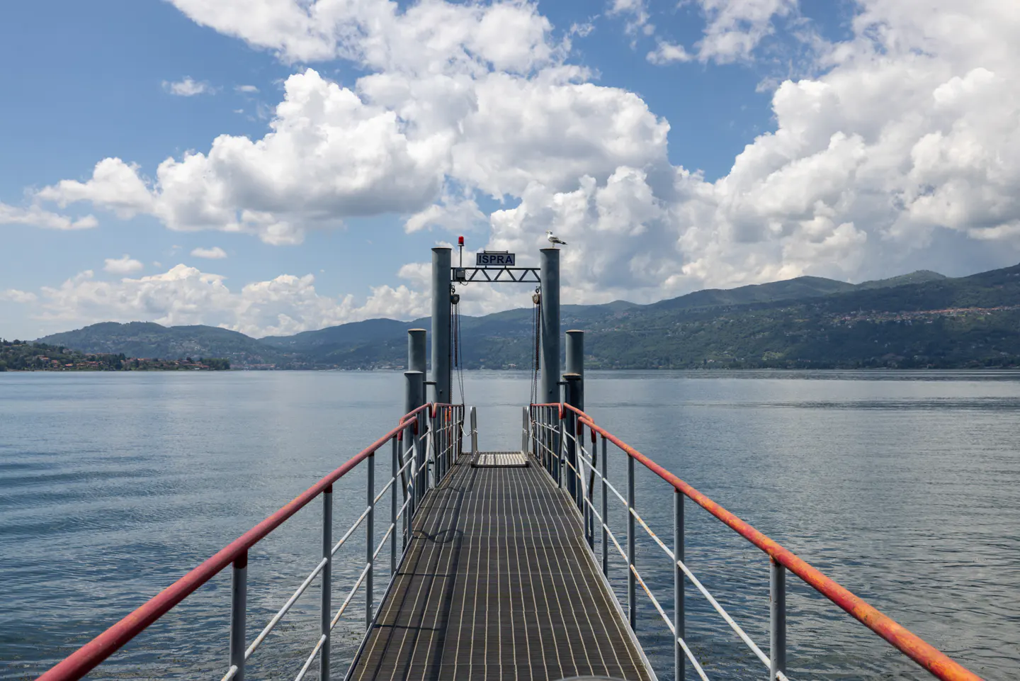 Pier with red railings leads to Lake Maggiore, Italy. Mountains and blue sky with clouds in the background.