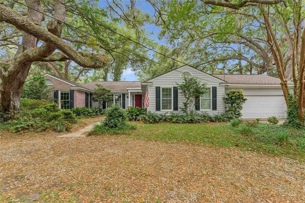 A single-story house with gray siding, black shutters, and a red door is surrounded by trees and greenery.