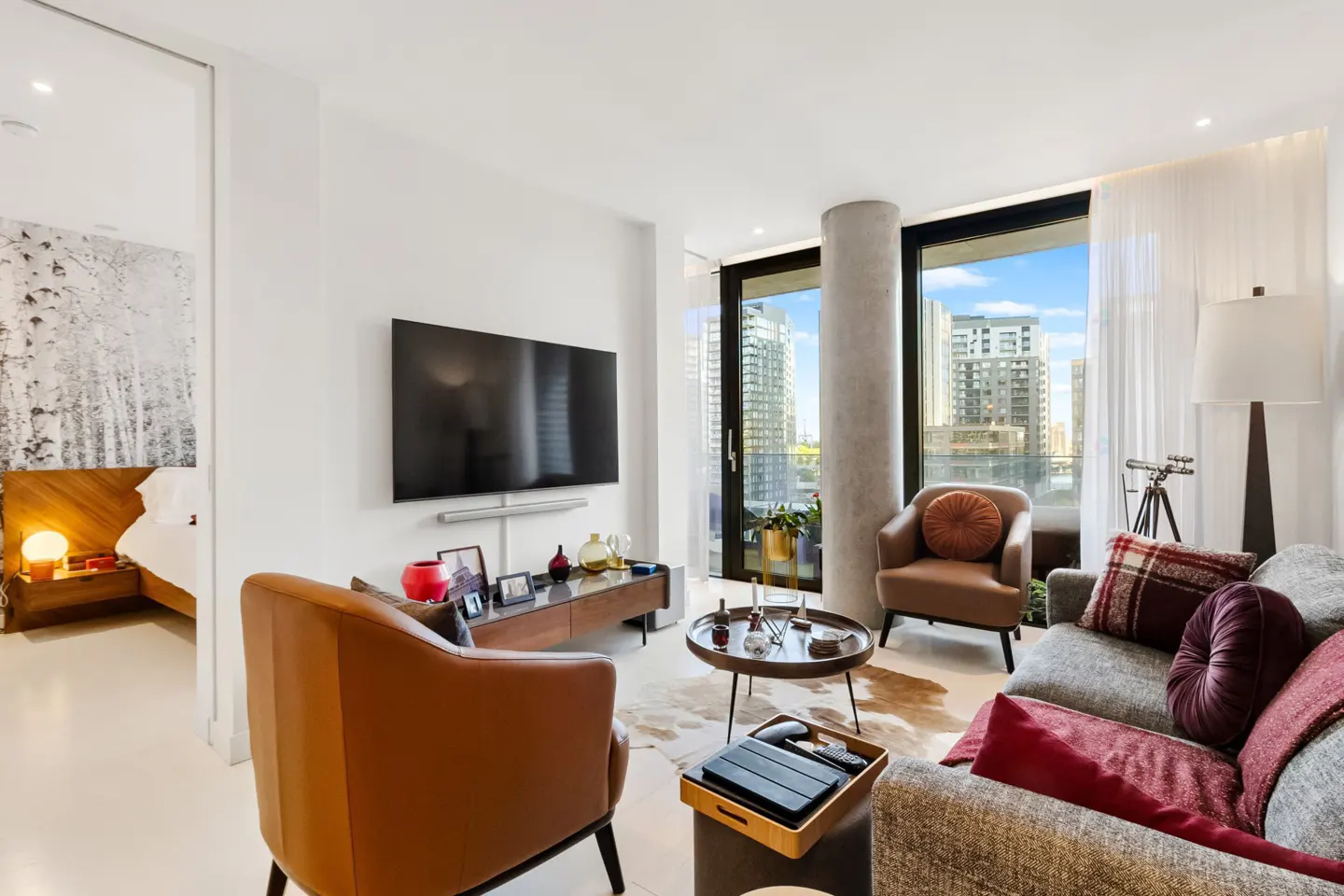 Bright living room with a TV, brown leather chairs, and a gray sofa. A bedroom is visible through an open doorway. City view through the window.