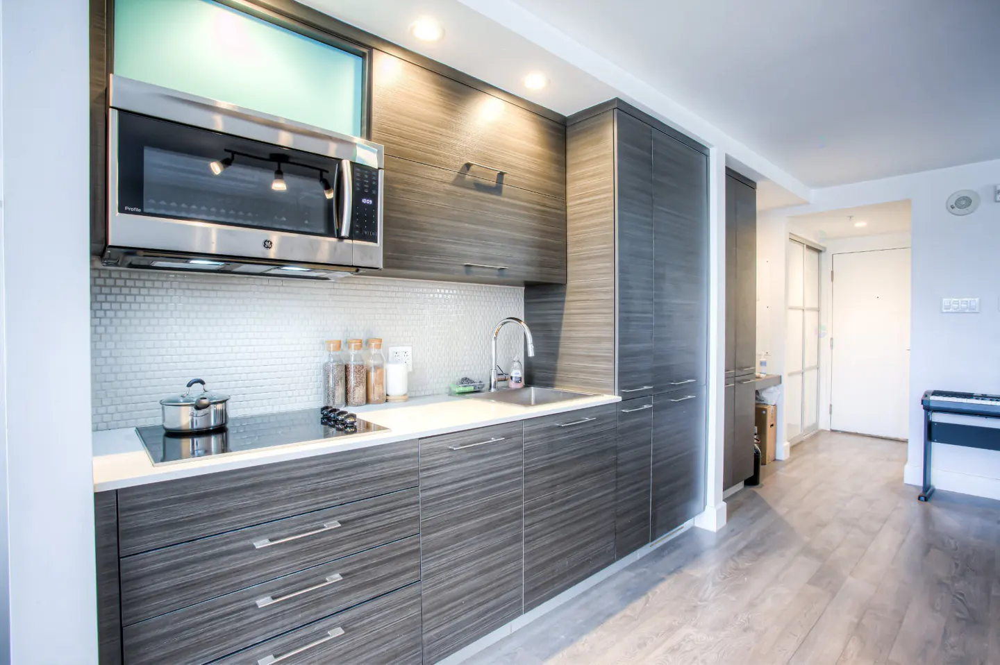 Modern kitchen with gray wood cabinets, stainless steel microwave, white countertop, and mosaic tile backsplash. Pot on stovetop.