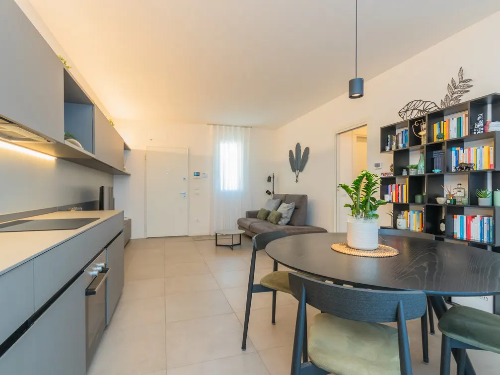 Modern open-plan apartment with a grey kitchen, round dining table, and a living area with a sofa and bookshelf.