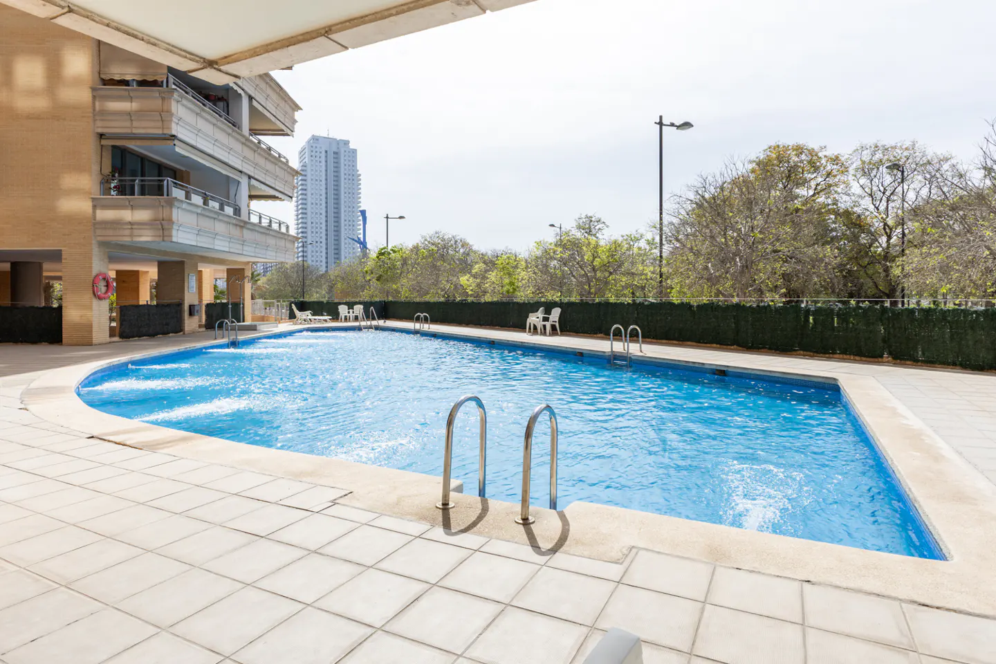 Outdoor pool with blue water, metal ladders, and tiled deck. Building and trees in the background.