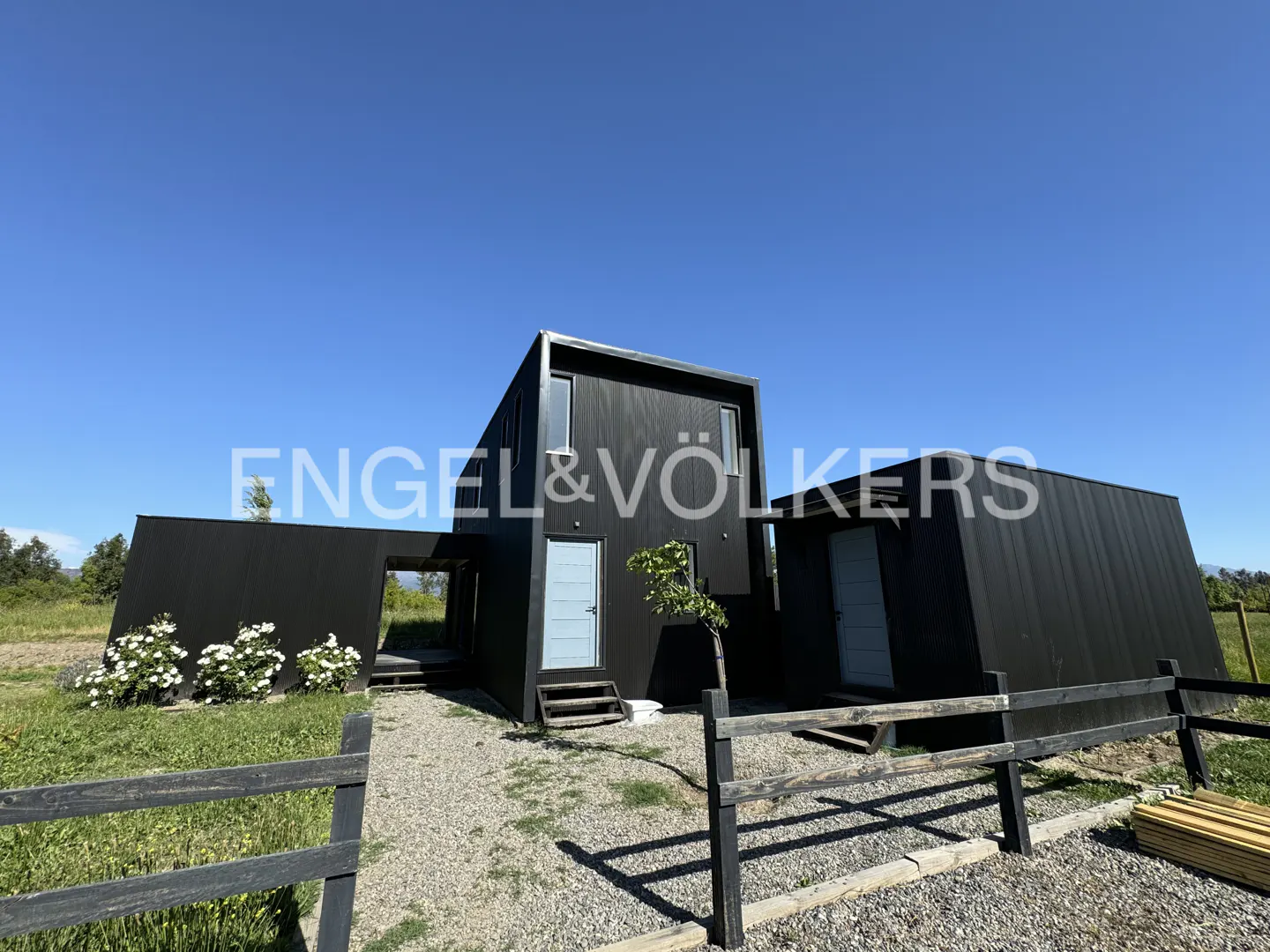 Modern black modular home with light blue doors, gravel yard, and wooden fence under a clear blue sky.