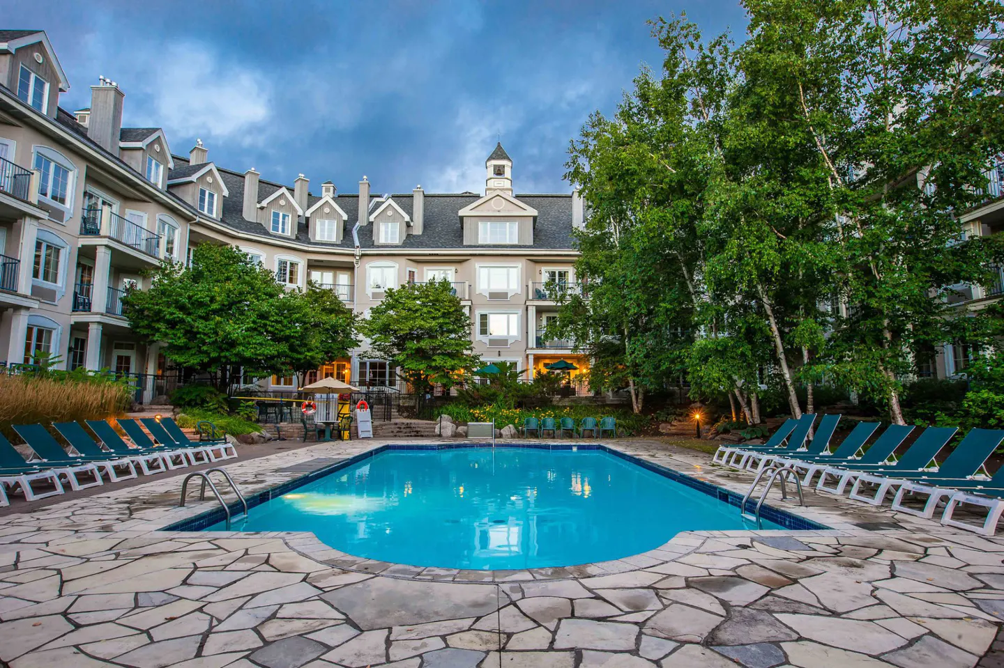 Outdoor pool with turquoise water, surrounded by stone patio, lounge chairs, and a multi-story building with trees.