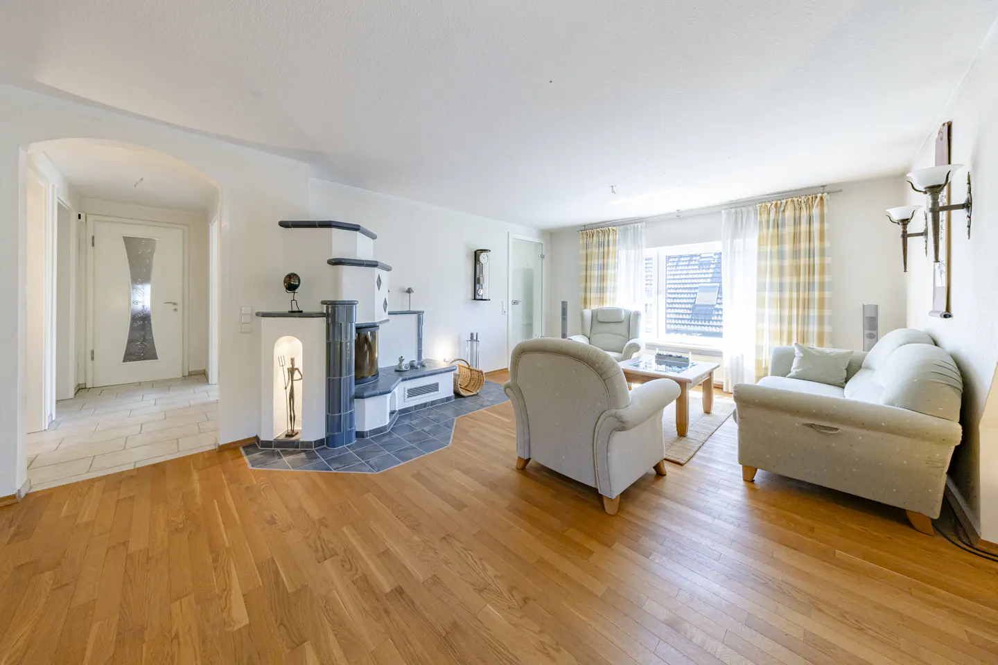 Bright living room with hardwood floors, a tiled fireplace, light-colored armchairs, a sofa, and a window with curtains.