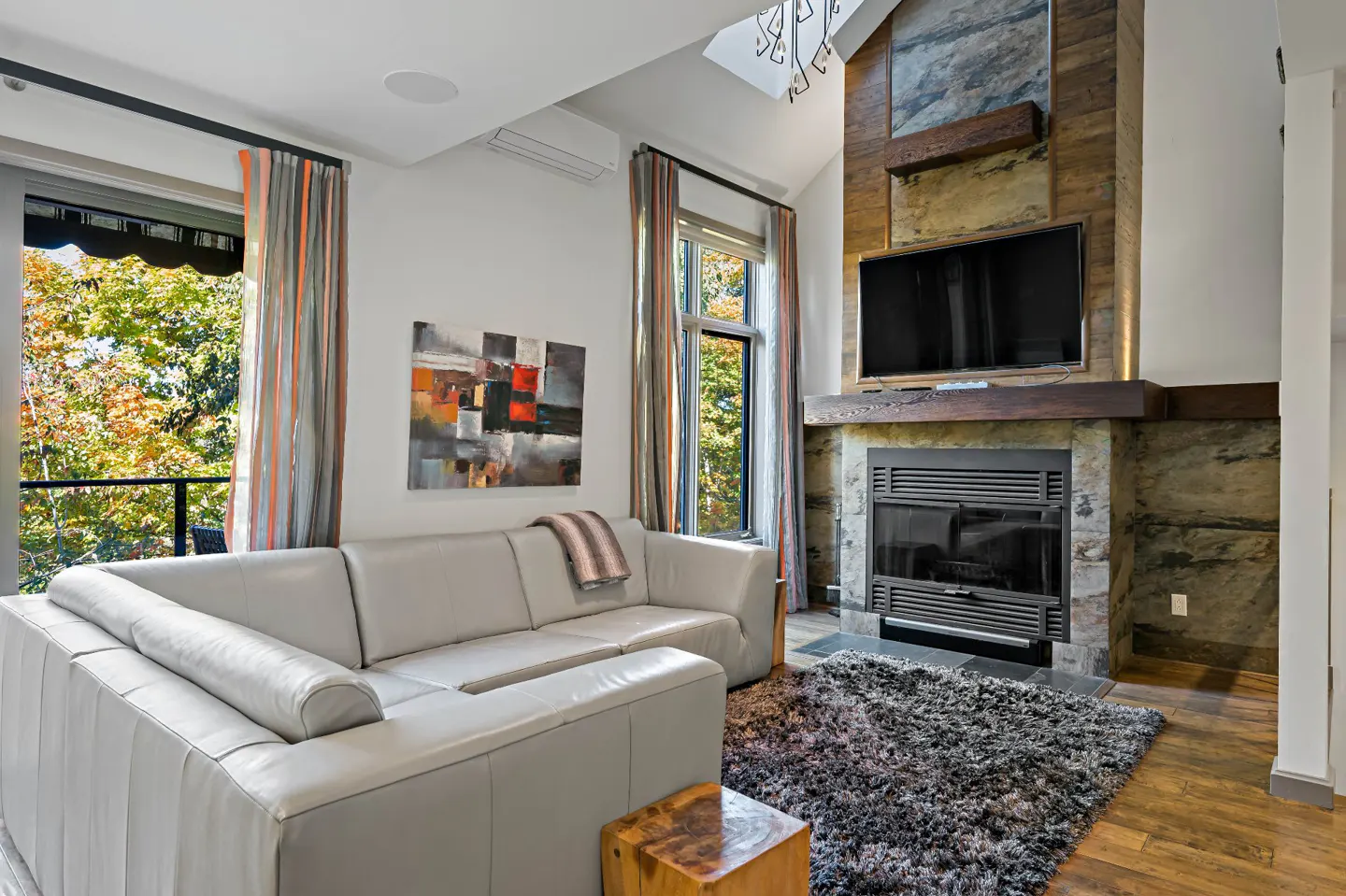 Living room with a gray leather sofa, stone fireplace with TV, and a view of colorful autumn trees.
