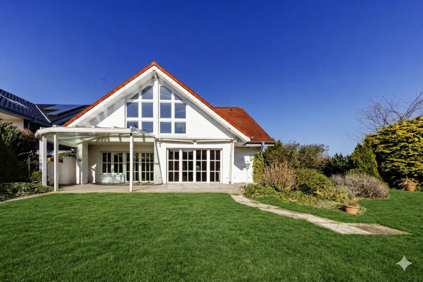 White house with red roof and large windows, surrounded by green lawn and garden under a clear blue sky.