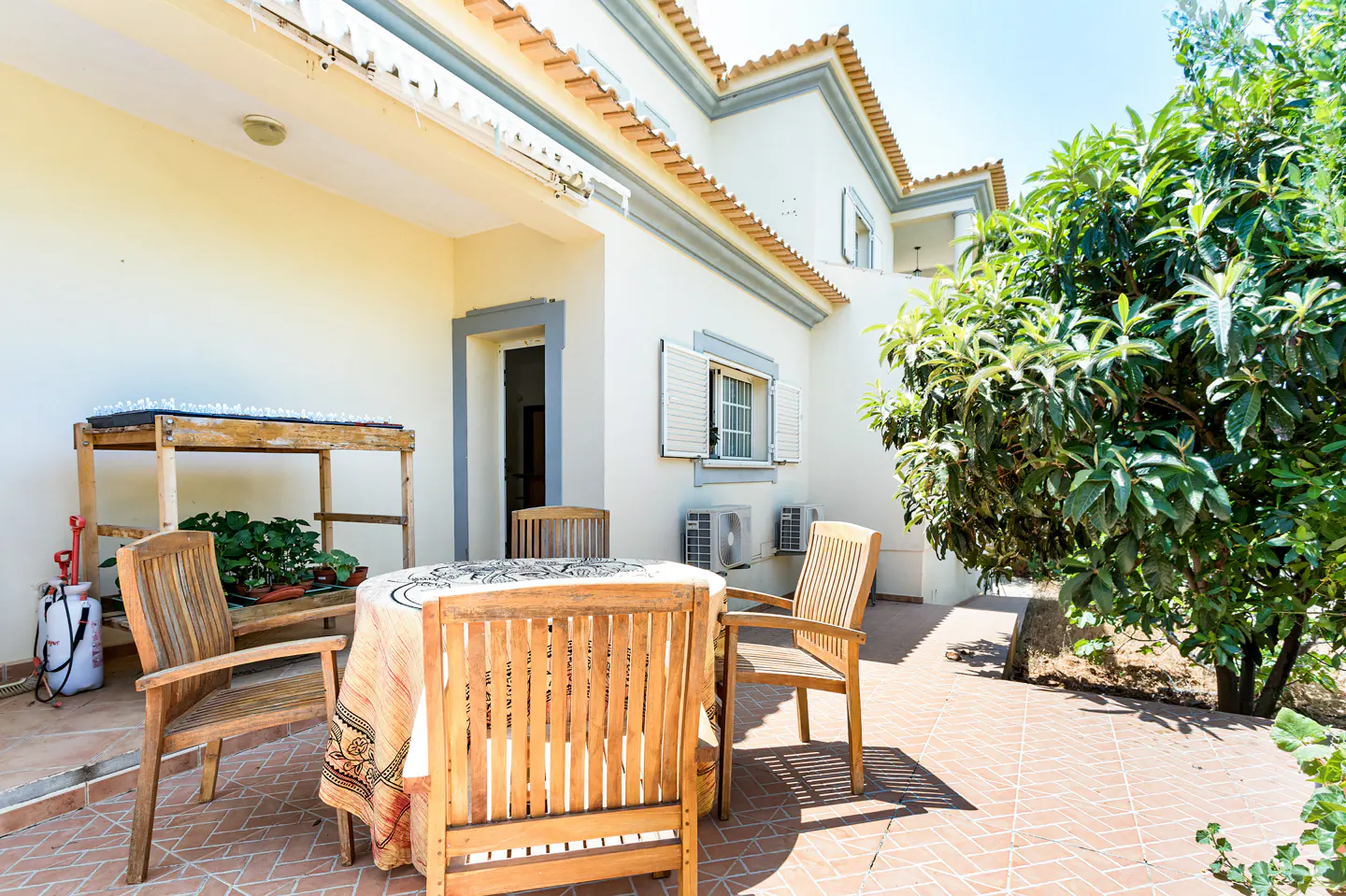 Outdoor patio with wooden table and chairs, patterned tablecloth, and potted plants. White building with orange roof tiles and green tree in background.