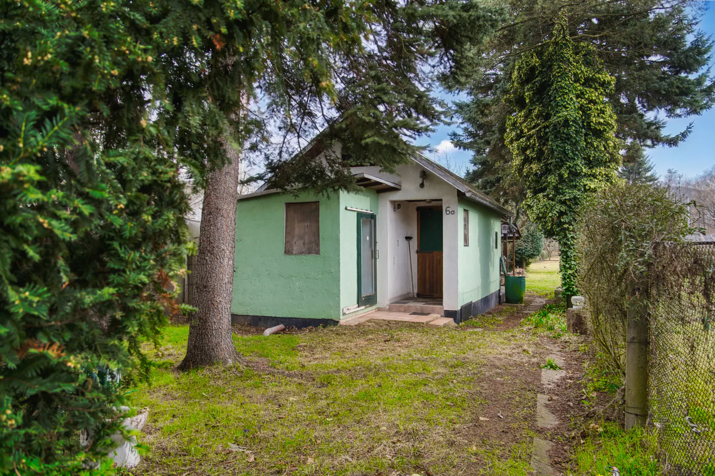 A small, one-story house with green and white walls, surrounded by trees and a grassy yard. The house has a brown door and a small window.