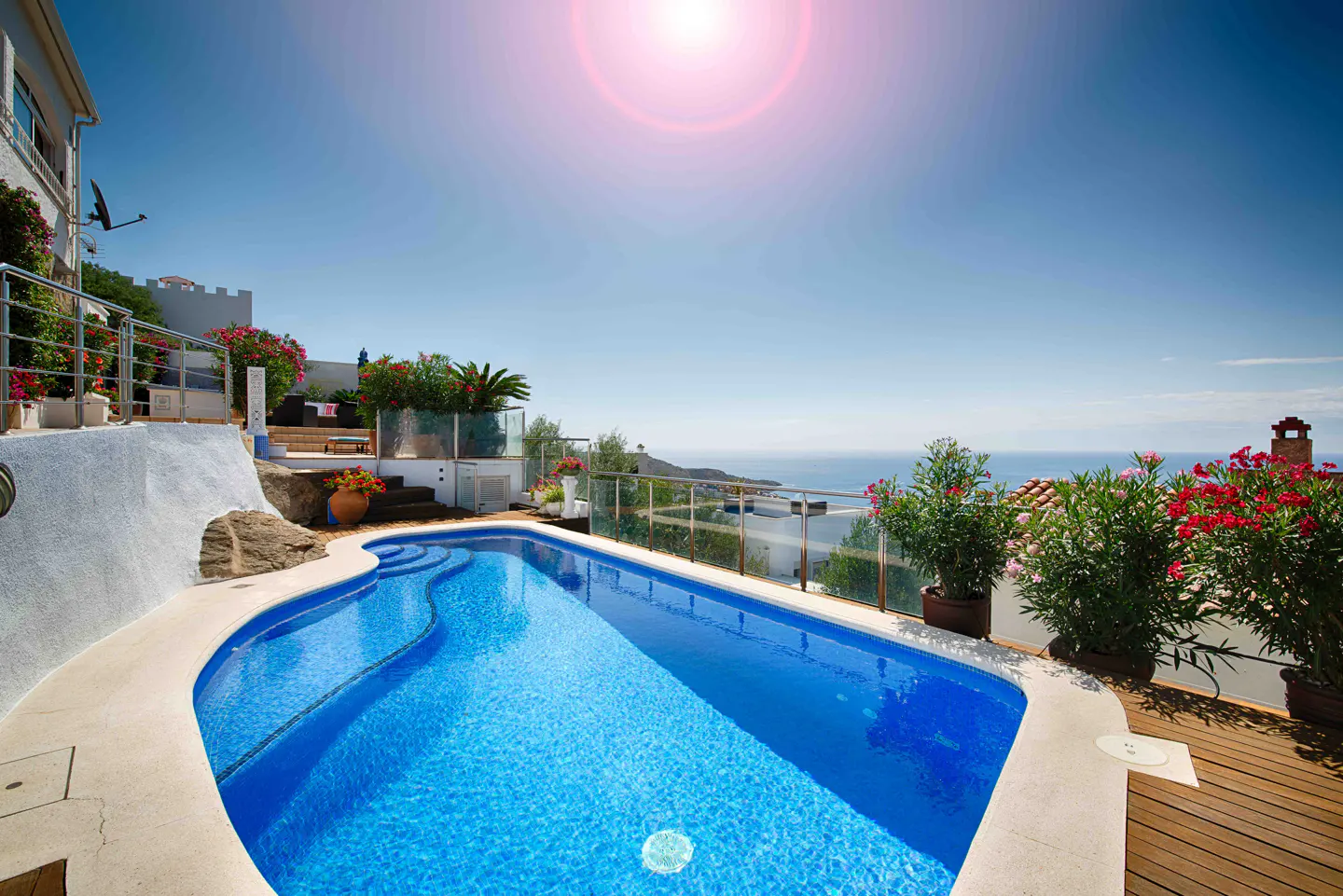 Bright blue pool with steps, surrounded by a white patio and wooden deck. Ocean view with clear blue sky and potted plants.